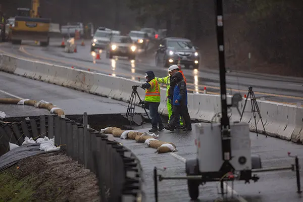 Workes in rptective gear using surveying equipment on a busy road