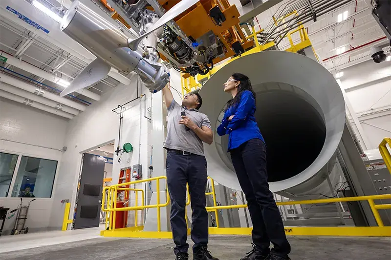 man and woman standing in front a a large tube shaped object while man reaches up to aircraft propeller