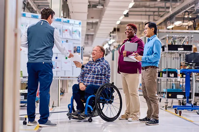 Man in business casual hands a packet of papers to man in wheel chair while woman and man look on within a testing environment