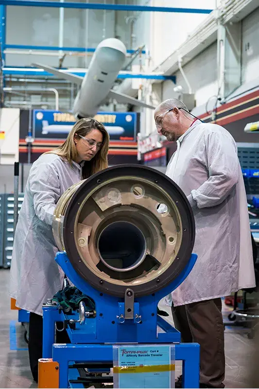 Woman and man in protective gear working on a large tubular object with a drone like craft hanging in the background