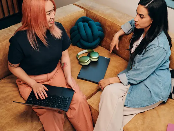 diverse women holding laptops face each other on a corner sofa talking chatting