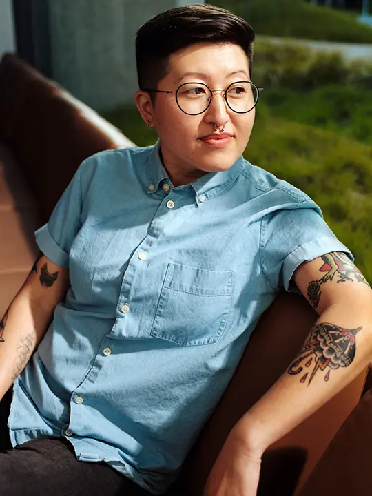 South Asian woman with short cropped hair and glasses sits in front of window of the green grass and shrubs