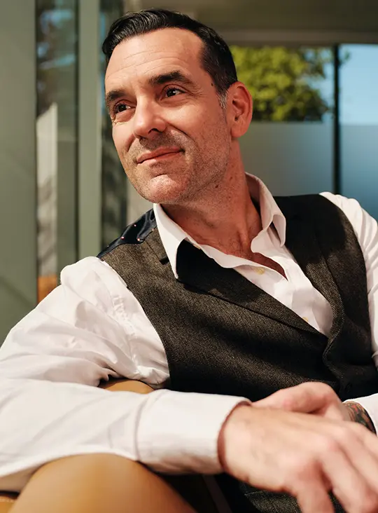 man in dress shirt and vest sits on sofa smiling looking out sunny window