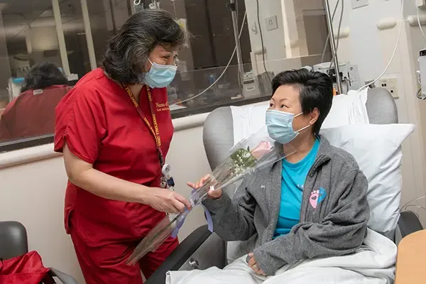 Nurse in surgical mask hands a long stem pink rose to a patien sitting in a chair propped up with a pillow