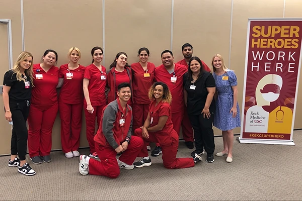 diverse men and women wearing red scrubs and name tags pose next to sign that reads super heroes work here