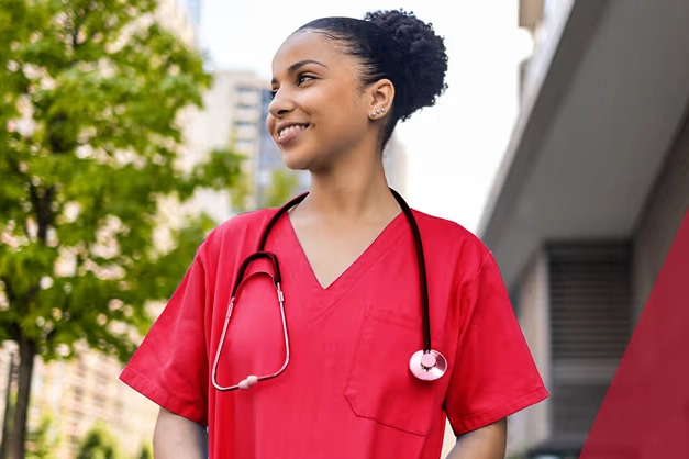 mixed race woman wearing scrubs and a stethoscope outdoors next to large building and Trees